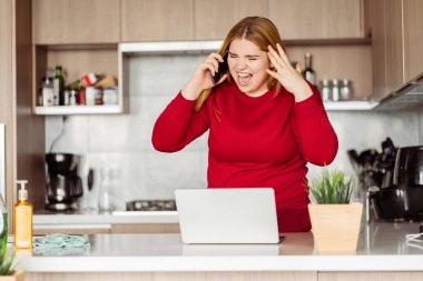 Üzgün, çekici, pozitif kadın cep telefonuyla konuşuyor, bağırıyor ve kızgın, dizüstü bilgisayar kullanıyor, evde uzaktan çalışıyor, oturma odasında duruyor. Kötü haber konsepti