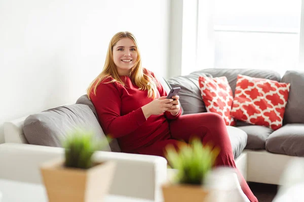 携帯電話を持っている魅力的な笑顔の白人ブロンドの女性 カメラを見ているアパートで快適なソファに座っているテキストメッセージ オンラインコミュニケーションの概念 — ストック写真