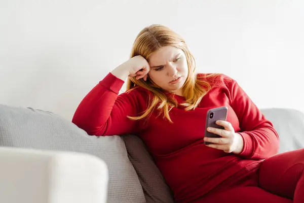 リビングルームでソファーに座っている携帯電話のテキストメッセージを保持している赤い服を着ている深刻な体の肯定的な女性 オンラインショッピングの概念 — ストック写真