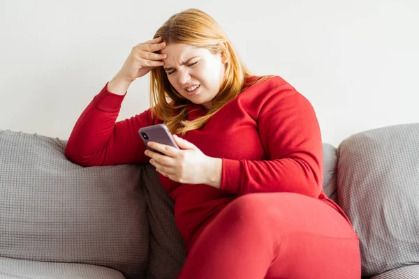 アパートのソファーに座って 携帯電話を使用して赤いカジュアルな服を着て 悪いニュース 悪いニュースをチェック ペンギン美しい若い女性 技術コンセプト — ストック写真