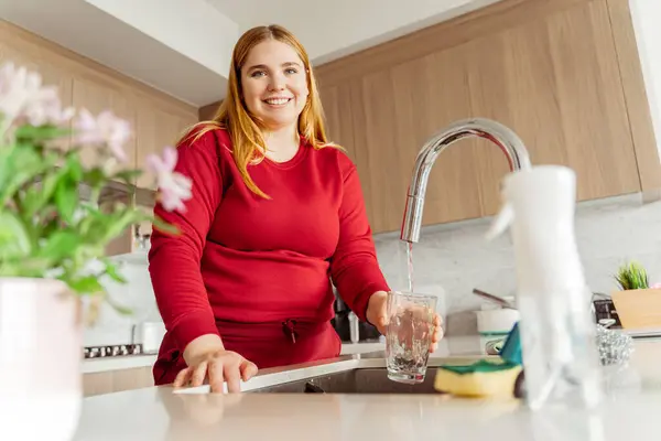 Usmívající Atraktivní Mladá Žena Červených Šatech Nalévá Vodu Kohoutku Kuchyni — Stock fotografie