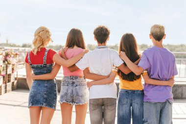 Arkadan bakan bir grup arkadaş, renkli tişörtler giyen çok kültürlü gençler sokakta dikilip birbirlerine bakıyorlar. Arkadaşlık kavramı 