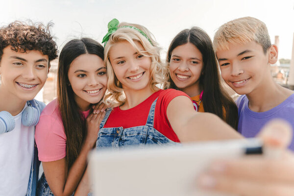 Group of smiling multiracial teenagers taking selfie, having video call on urban street. Young blogger influencer recording video standing together with friends. Technology. Positive lifestyle concept
