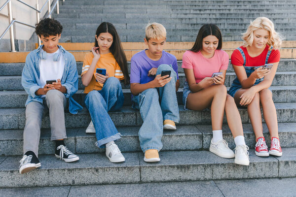 Группа серьезных друзей, многорасовые подростки, держащие мобильные телефоны, смотрящие видео, чаты, общение онлайн сидя на лестнице. Технологическая зависимость, концепция социальных сетей 