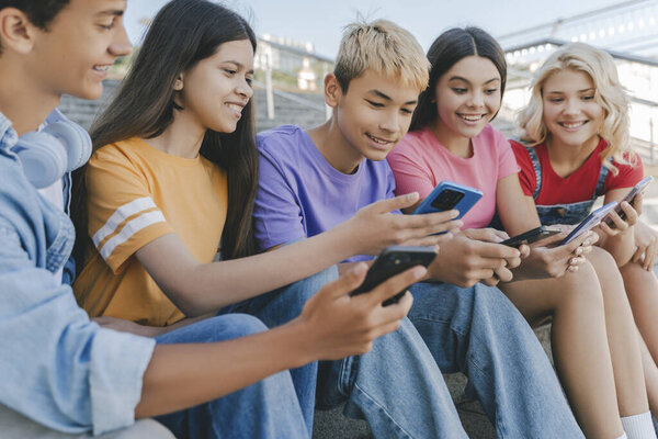 Group of smiling friends, multiracial teenagers holding mobile phones watching video, communication online, playing mobile game, talking sitting on stairs. Technology, social media concept