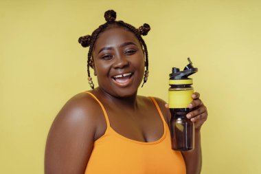 Güzel Afrikalı Amerikalı kadın elinde şişeyle sarı arka planda izole edilmiş kameraya bakıyor. Sağlık hizmeti konsepti, artı boyut modeli
