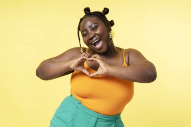 Gülümseyen neşeli vücut pozitif Afrikalı kadın, sarı arka planda elleri izole bir kalp gösteriyor. Artı Nijerya modeli rahatlama, doğal güzellik konsepti