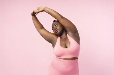 Pembe spor sutyen giyen ve pembe arka planda izole edilmiş egzersizler yapan güzel, modern ve iri yarı bir kadın. Vücut pozitif kadın modeli, sağlık konsepti, motivasyon konsepti