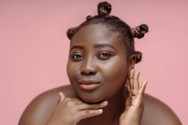 Aynaya bakan büyüleyici Afrikalı kadın pembe arka planda izole edilmiş tenine dokunuyor. Vücut pozitif kavram, cilt bakımı