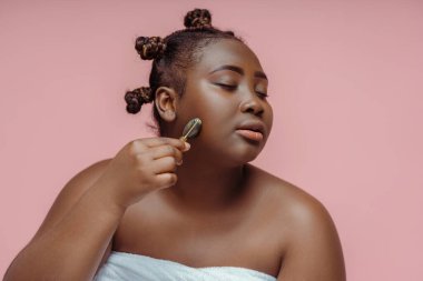 Güzel, gözleri kapalı, pembe arka planda yüz masajı yapan Afrikalı bir kadın. Güzellik prosedürleri konsepti