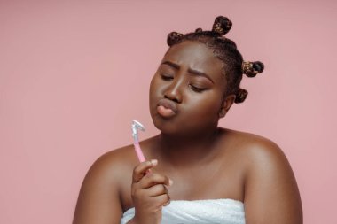 Komik, neşeli Afrikalı kadın pembe arka planda izole edilmiş havluyla duş aldıktan sonra jilet tutuyor. Çekici kadın sabah prosedürleri, cilt bakımı