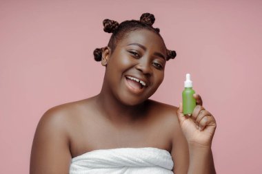 Havluyla duş alan neşeli Afro-Amerikalı kadın. Pembe arka planda yüz yağını tutan şık bir saç stili. Güzel bayan kameraya bakıyor, poz veriyor.