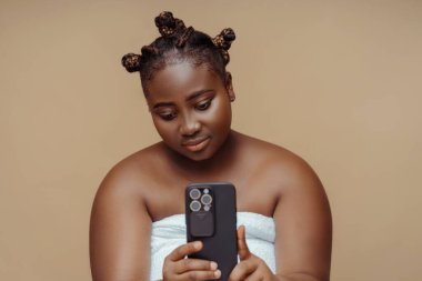 Havlu giymiş, elinde cep telefonu olan çekici Afro-Amerikan kadın aynaya bakarak selfie çekiyor. Vücut ve cilt bakımı kavramı