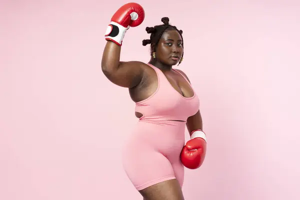 stock image Pensive beautiful Nigerian woman wearing sports uniform red boxing gloves looking at camera isolated on pink background. Authentic body positive African model posing for picture, copy space