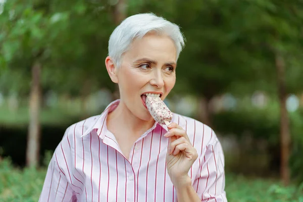 快乐而时尚的灰白头发女人吃着美味的冰淇淋 站在公园里遥望远方 积极的生活方式概念 — 图库照片