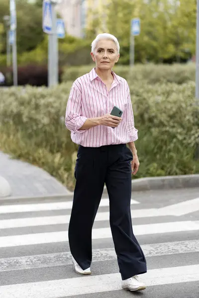 Серьёзная Пожилая Женщина Повседневной Одежде Держит Мобильный Телефон Идущий Улице — стоковое фото
