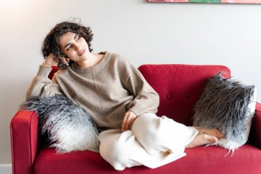 Romantik ortadoğulu kadın kırmızı kanepede dinlenirken gözlerini kaçırıyor. Güzel gülüşü olan genç kıvırcık bayan evde oturuyor, haftasonunu yalnız geçiriyor.