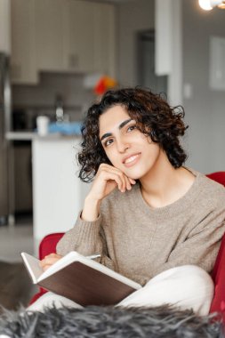 Kıvırcık kadın kameraya bakıyor, kırmızı koltuktaki mandıra defterine notlar yazıyor. Güzel romantik Orta Doğulu kadın evde.