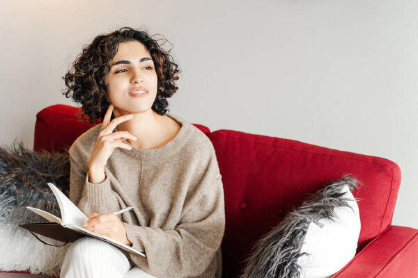 Smiling positive curly woman sitting at red sofa, making notes in notebook at home. Attractive middle eastern female writer in casual clothes. Project planning, online education 