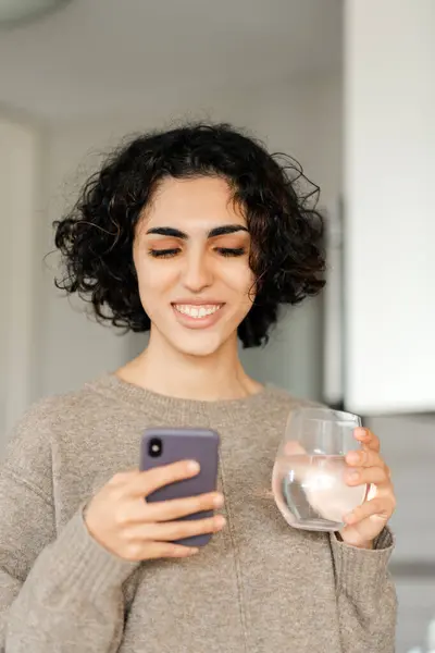 Smiling curly woman holding smartphone and glass of water, watching videos at home. Happy modern female using mobile app shopping online, ordering food, reading text message, check email