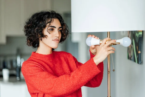 Curly middle eastern woman independently changes burned-out light bulb in order to achieve high-quality lighting in house. Girl with smile inserts light bulb into chandelier