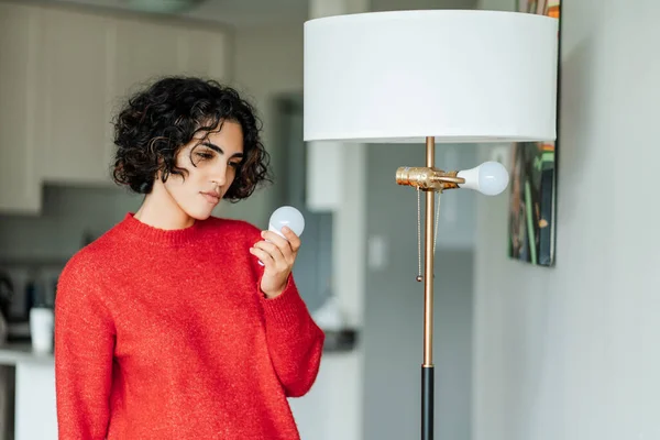 Curly middle eastern woman holding light bulb for chandelier and looking on it at home. Girl independently changes burned-out light bulb in order to achieve high-quality lighting in house