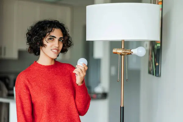 Smiling middle eastern woman holding light bulb for chandelier and looking at camera at home. Girl changes burned-out light bulb in order to achieve high-quality lighting in house