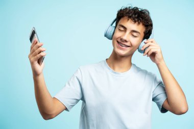 Kulaklık takan gülümseyen genç bir çocuğun portresi. Müzik dinliyor, cep telefonunu mavi arka planda kapalı tutuyor. Teknoloji konsepti
