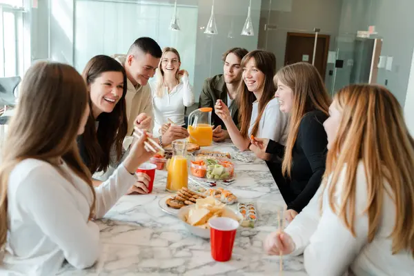 Cheerful friends gathering at dining table with juice and sushi. Happy young people eating, celebrating holiday, talking, relaxing together on weekend