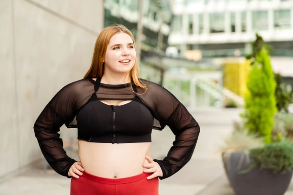 Happy lady in sports top and leggings posing and looking to the side in the city. Body positive, plus size, motivation