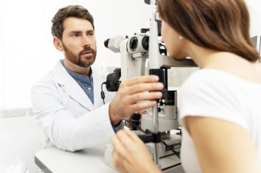 Erkek optometrist, kadın hastanın gözünü incelerken dürbün lamba kullanıyor. Tıbbi muayene