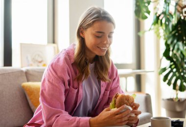 Güzel bir kadının portresi şık günlük giysiler içinde elinde burger, kanepede otururken yemek yemek, öğle yemeği. Beslenme kavramı