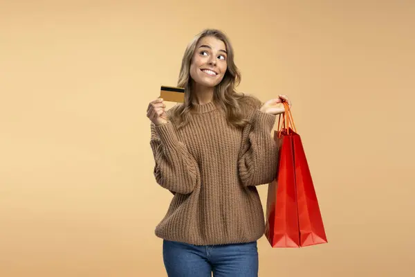 魅力的な笑顔の女性 クレジットカードと赤いショッピングバッグを持ち ベージュの背景に隔離された服を選択し スペースをコピーします ショッピング セール ブラックフライデー 広告コンセプト — ストック写真