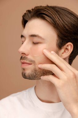 Hidrojel göz bandı sürerken kameraya poz veren genç bir adam. Bej arka planda izole edilmiş. Cilt bakımı, kozmetik, kaplıca tedavisi