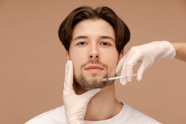 Eldiven takan doktor elleri, adamın yüzünde şırınga yapıyor. Bej arka planda izole edilmiş. Güzellik bakımı, yaşlanma karşıtı prosedürler, estetik ameliyat konsepti.