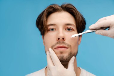 Hasta erkek yüzünün yanında neşter tutan doktor elleri. Kozmetik Operasyonu. Mavi arka planda izole edilmiş. Güzellik bakımı, yaşlanma karşıtı prosedürler, estetik ameliyat konsepti.