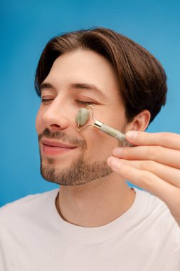 Mutlu adamın yüzüne yeşim taşıyla masaj yapıp stüdyoda poz verişini yakından çek. Mavi arka planda izole edilmiş. Cilt bakımı, güzellik, şişkinliği azaltma, yaşlanmayı önleme yöntemi