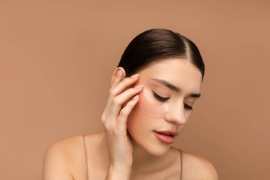 Hidrojel göz bantlarıyla poz veren güzel genç bayan portresi. Bej arka planda izole edilmiş. Cilt bakımı, doğal güzellik, kaplıca tedavisi