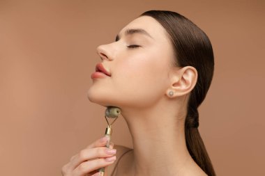 Çekici genç bir kadının portresi yeşim taşı yüz silindiri kullanıyor ve gözleri kapalı gülümsüyor. Bej arka planda izole edilmiş. Cilt bakımı, yaşlanma önleme prosedürü