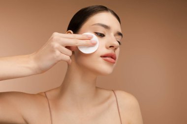 Yüzü temizlerken pamuk süngerle makyaj yapan genç bayanın portresi, günlük sağlıklı güzellik prosedürü. Bej arka planda izole edilmiş. Mükemmel kozmetoloji cilt bakımı