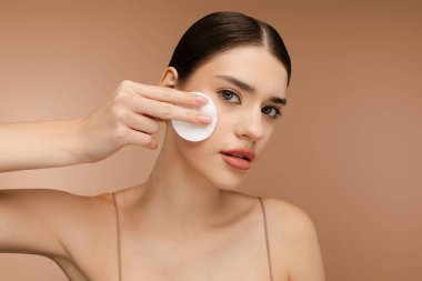 Yüzünü temizlerken, kameraya bakarken pamuk sünger kullanan güzel bir kadının portresi. Bej arka planda izole edilmiş. Mükemmel kozmetoloji cilt bakımı