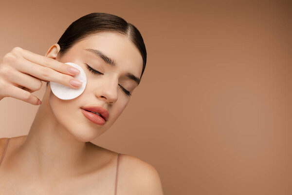 Портрет красивой брюнетки, использующей губку и удаляющей макияж с лица. Принято. Изолированный на бежевом фоне. Идеальный уход за кожей