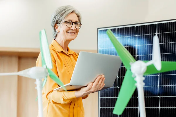 Smiling senior woman, engineer, developer using laptop standing near solar panel and windmill working in modern office. Alternative, wind energy, innovation concept