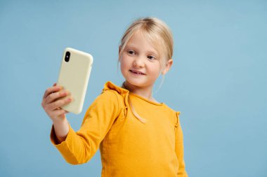 Güzel, gülümseyen küçük bir kızın portresi, cep telefonu tutan, selfie çeken, video kaydeden, sosyal medya, mavi arka planda izole edilmiş poz veren. Teknoloji konsepti