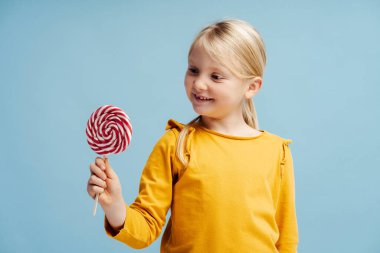 Sıradan kıyafetler giymiş, elinde lolipop şekeri tutan, mavi arka planda izole bir şekilde duran sevimli, gülümseyen küçük bir kızın portresi. Tatlı ve yiyecek kavramı.