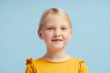 Sevimli, güzel, güler yüzlü, sıradan elbiseli küçük bir kızın portresi, mavi arka planda izole bir şekilde duran kameraya bakıyor. Çocukluk konsepti, neşeli çocuk.