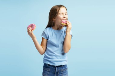 Çekici, gülümseyen, günlük kıyafetler giyen, lezzetli pembe çörekler yiyen, mavi arka planda tek başına duran küçük bir kızın portresi. Yemek kavramı, tatlı.