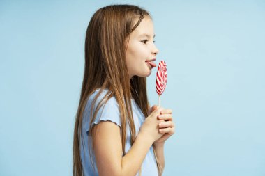 Sıradan kıyafetler giyen, lolipop şekeri tutan, ona bakan, yemek yiyen, mavi arka planda tek başına duran pozitif küçük bir kızın portresi. Tatlı ve yiyecek kavramı.