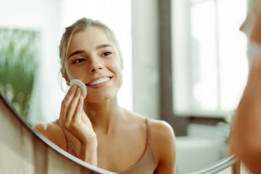 Genç, gülümseyen bir kadın makyaj malzemelerini pamukla yıkıyor aynaya bakıyor, banyoda duruyor. Cilt bakımı, tedavi kavramı