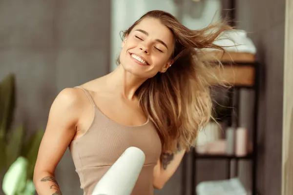 Happy beautiful young woman drying her hair with dryer, wearing tank top standing in bathroom. Concept of hair care, hairstyle